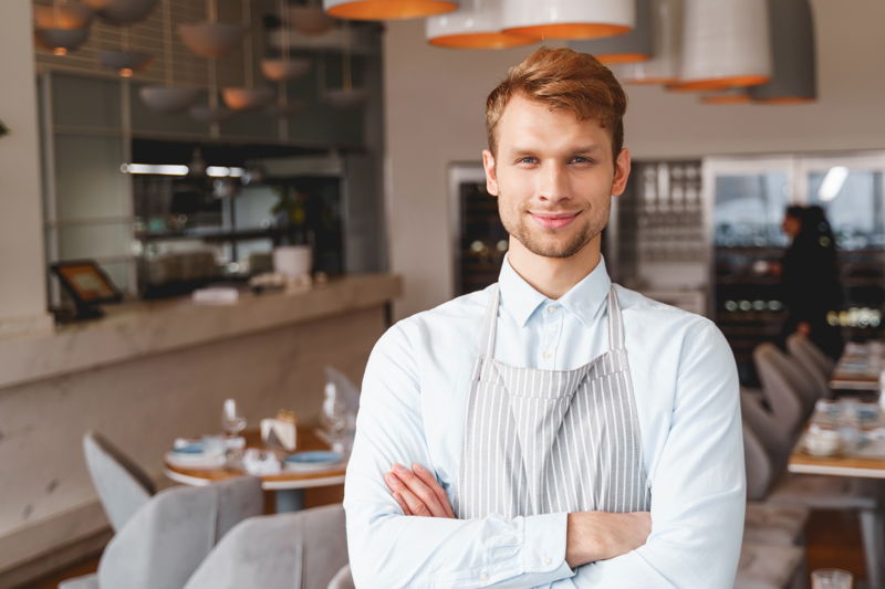 datenschutz gastronomie Ein junger Geschäftsführer mit Servirschürze steht mit verschränkten Armen in einem Restaurant.